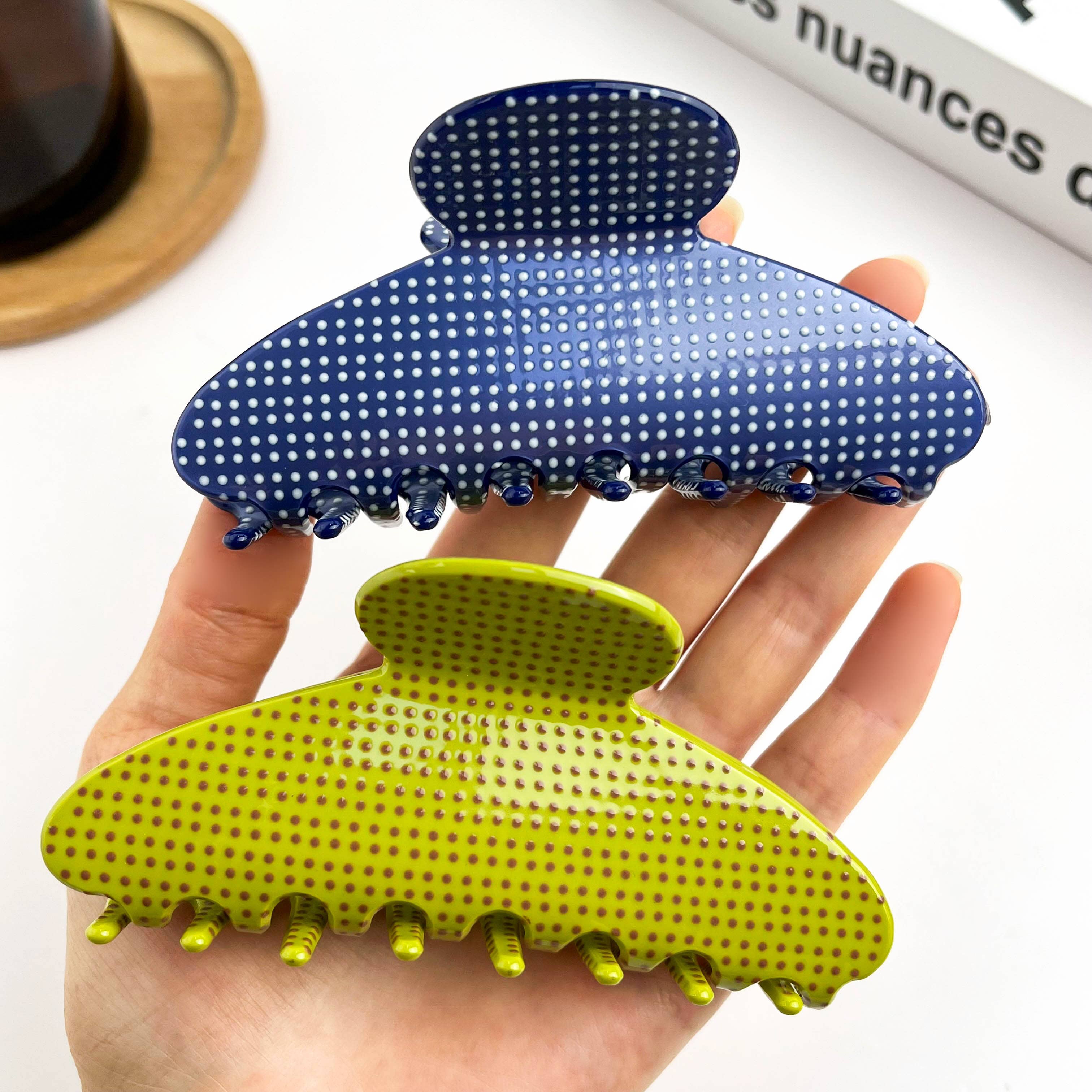 Hand holding two polka dot hair clips, one blue and one green, on a white background.