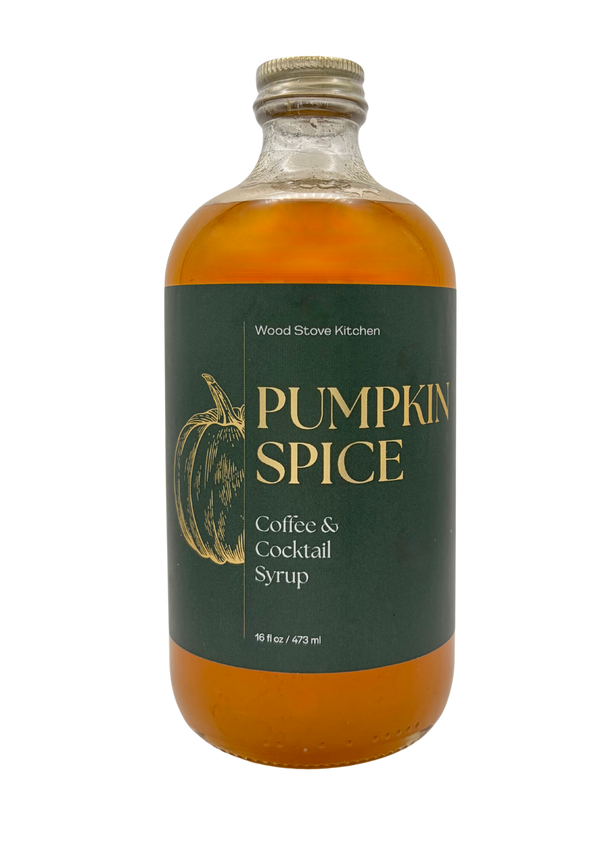 A bottle of Wood Stove Kitchen Pumpkin Spice Coffee & Cocktail Syrup, featuring an amber-colored liquid in a clear glass bottle with a dark green label and gold lettering.