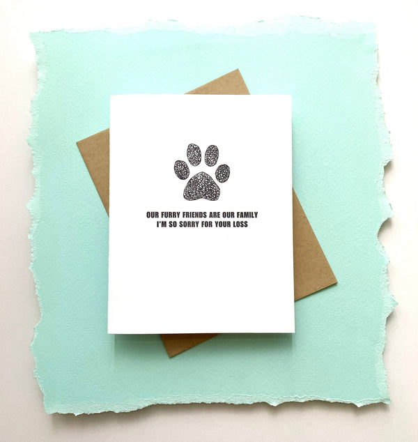 A sympathy card with a white background featuring a black paw print and the text 'Our Furry Friends Are Our Family I'm So Sorry For Your Loss' letterpressed on it, accompanied by a recycled envelope.