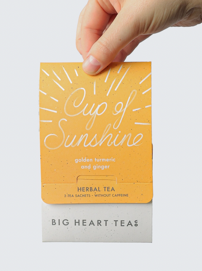 A hand holding a tea bag packaging labeled 'Cup of Sunshine' by Big Heart Teas, with sunBeam design in yellow color.